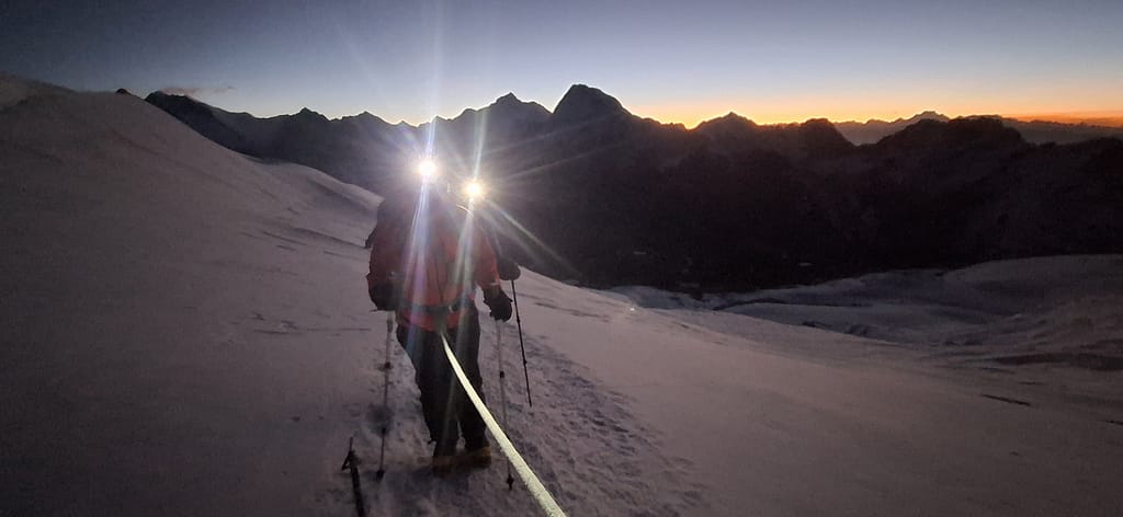 Zifir karanlıkta yürüyüş yapan bir dağcı, başında kafa lambası var, çevresinde dağ silüetleri ve sabahın ilk ışıkları gözlemleniyor.