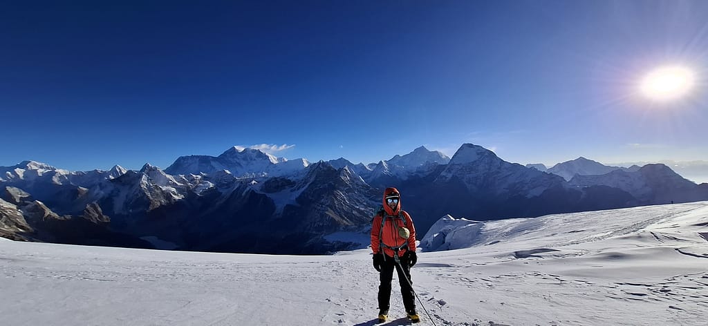 Bir dağcı, yüksek irtifada kar kaplı bir zeminde duruyor; arka planda etkileyici dağ manzaraları ve açık bir mavi gökyüzü var.