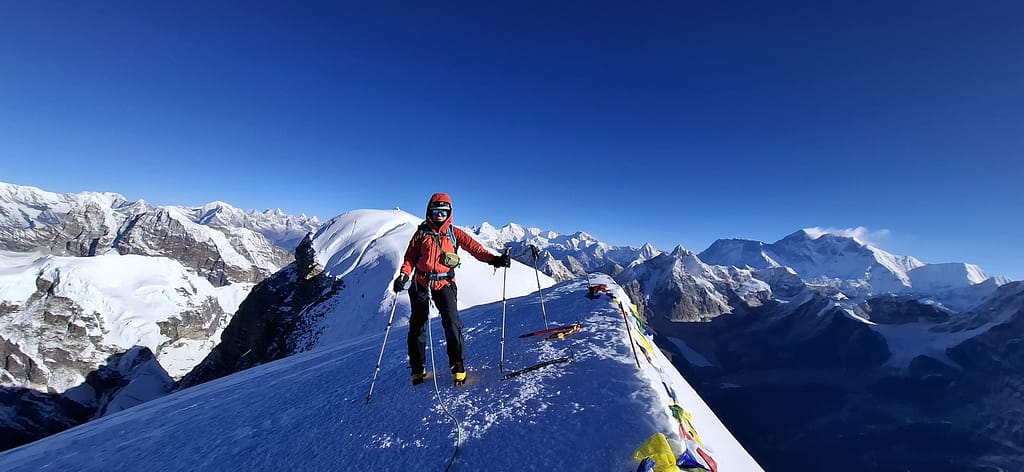 Bir dağ zirvesinde bir dağcı, arka planda görkemli beyaz dağlarla dolu bir manzara eşliğinde duruyor. Kişi, dağcılık ekipmanlarıyla donanmış ve renkli bir kıyafet giymiş.