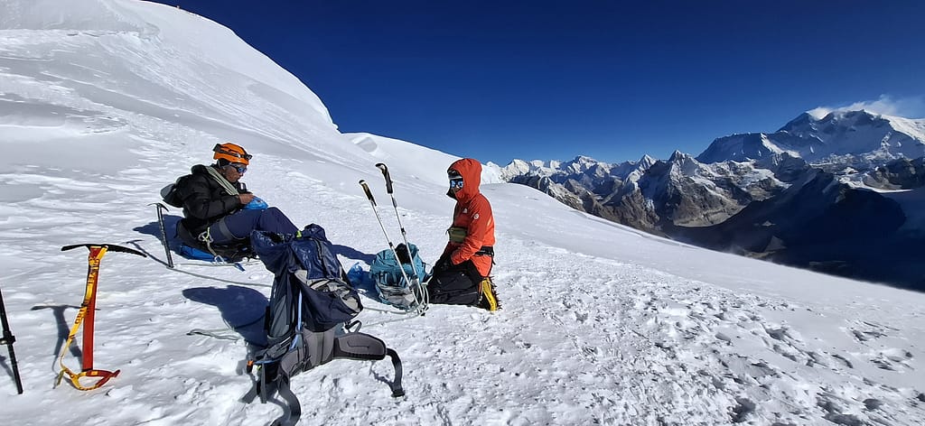 İki dağcı, yüksek irtifada, buzlu bir zeminde oturarak dinleniyor. Etrafta karla kaplı dağlar manzarası ön planda.