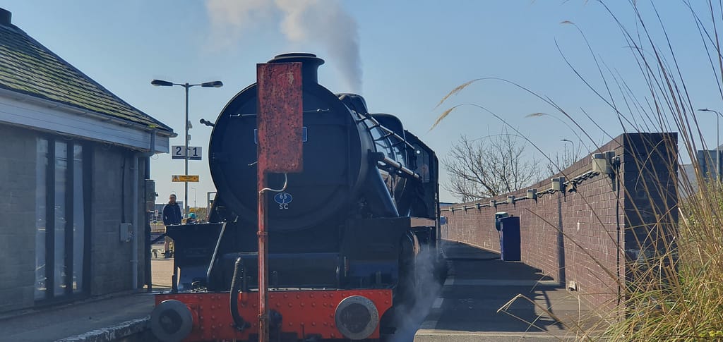 Eski bir buharlı lokomotif trenin arka görünümü, yanındaki tabelada 211 yazıyor. Güneşli bir günde durakta bekleyen tren, hafif buhar çıkartıyor.