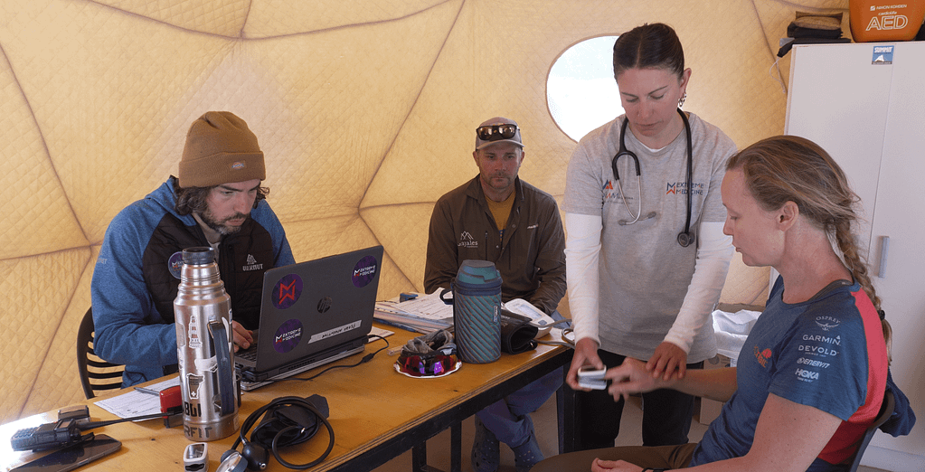 Bir tıbbi kontrol sırasında hemşire, sağlık ekipmanlarını kullanarak hastanın durumunu değerlendiriyor. Diğer iki kişi bilgisayarda çalışıyor.