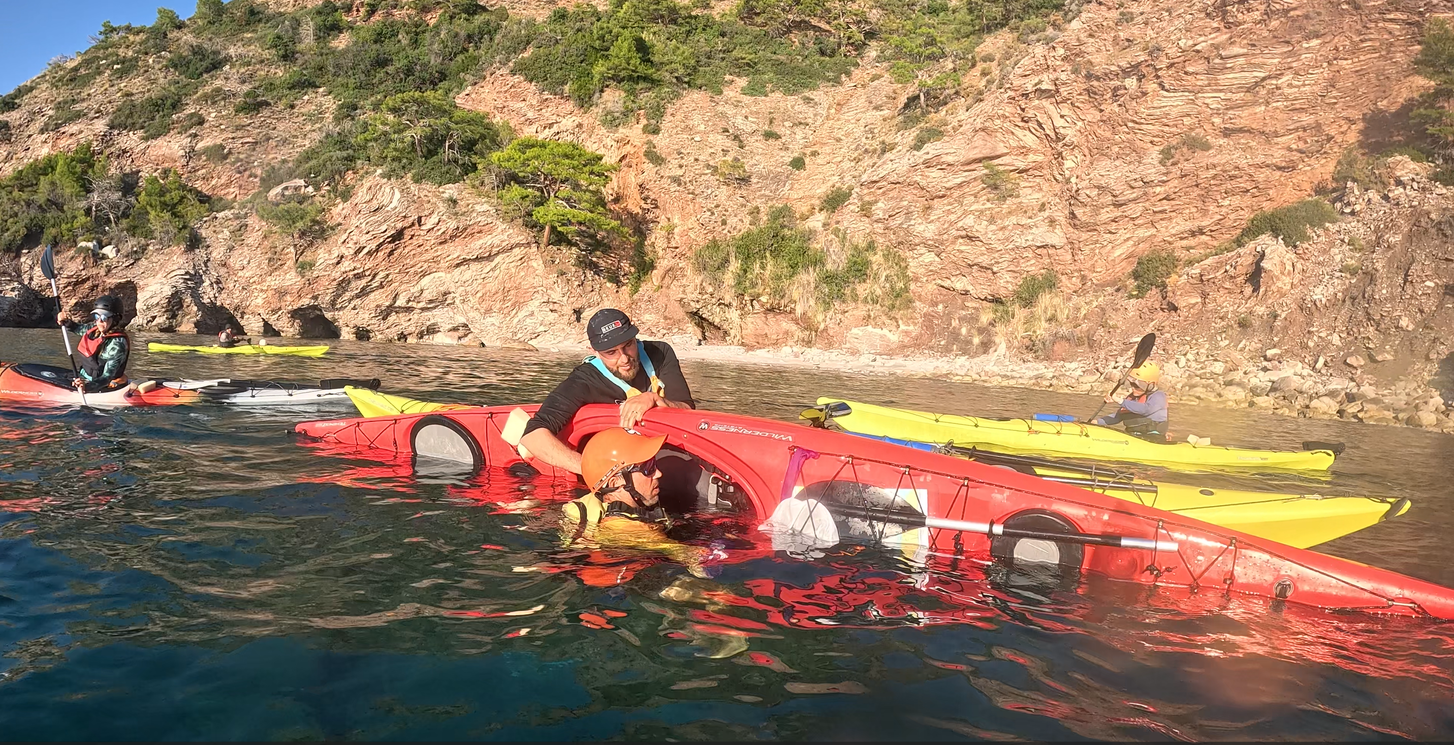 Deniz kayağı eğitimi sırasında bir kişinin kayıkta self-rescue teknikleri uyguladığı ve diğer katılımcıların yardım ettiği bir sahne.
