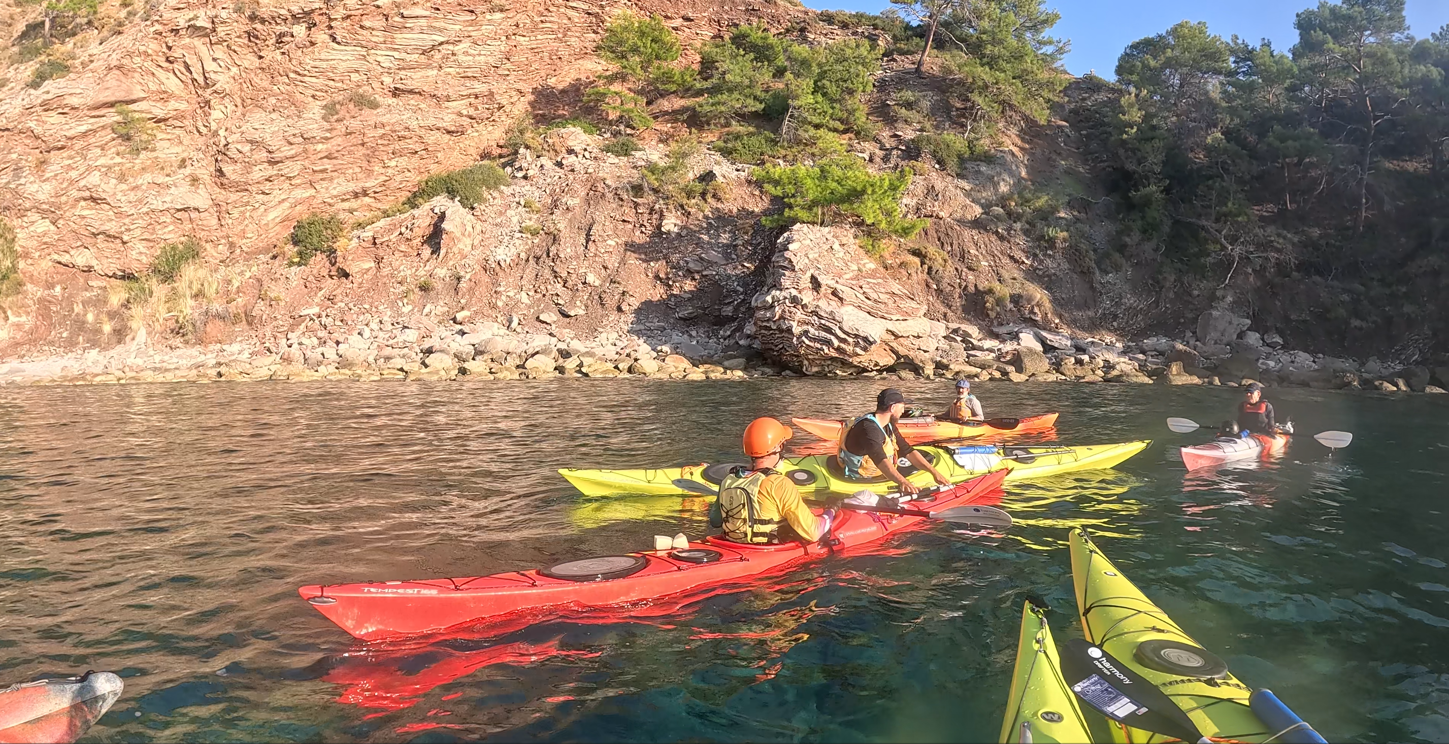 Bir grup kayakçının, deniz kayıklarıyla kayalık bir kıyıya yakın konumlandığı bir manzara.