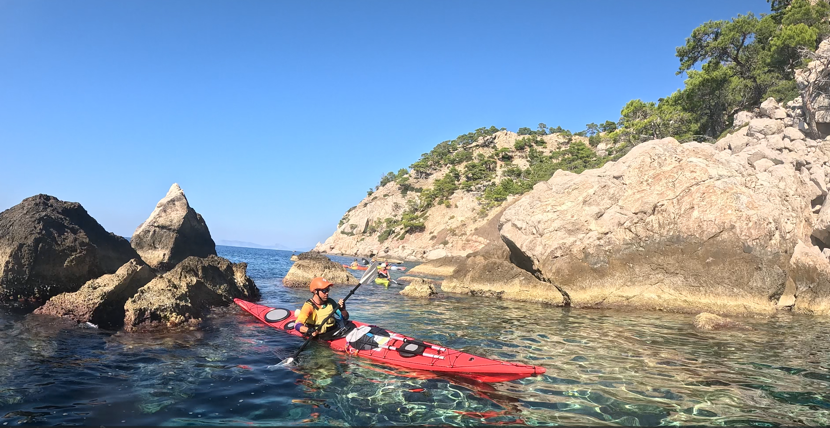 Bir deniz kayığı üzerindeki kişi, kayalık bir kıyı boyunca ilerliyor. Suyun berraklığı ve çevredeki doğal manzara göz önünde. Arkada başka deniz kayakçıları yer alıyor.