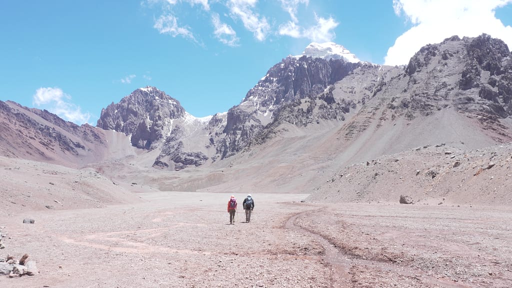 İki dağcı, kuru bir vadide, sarp dağların önünde yürüyüş yapıyor.