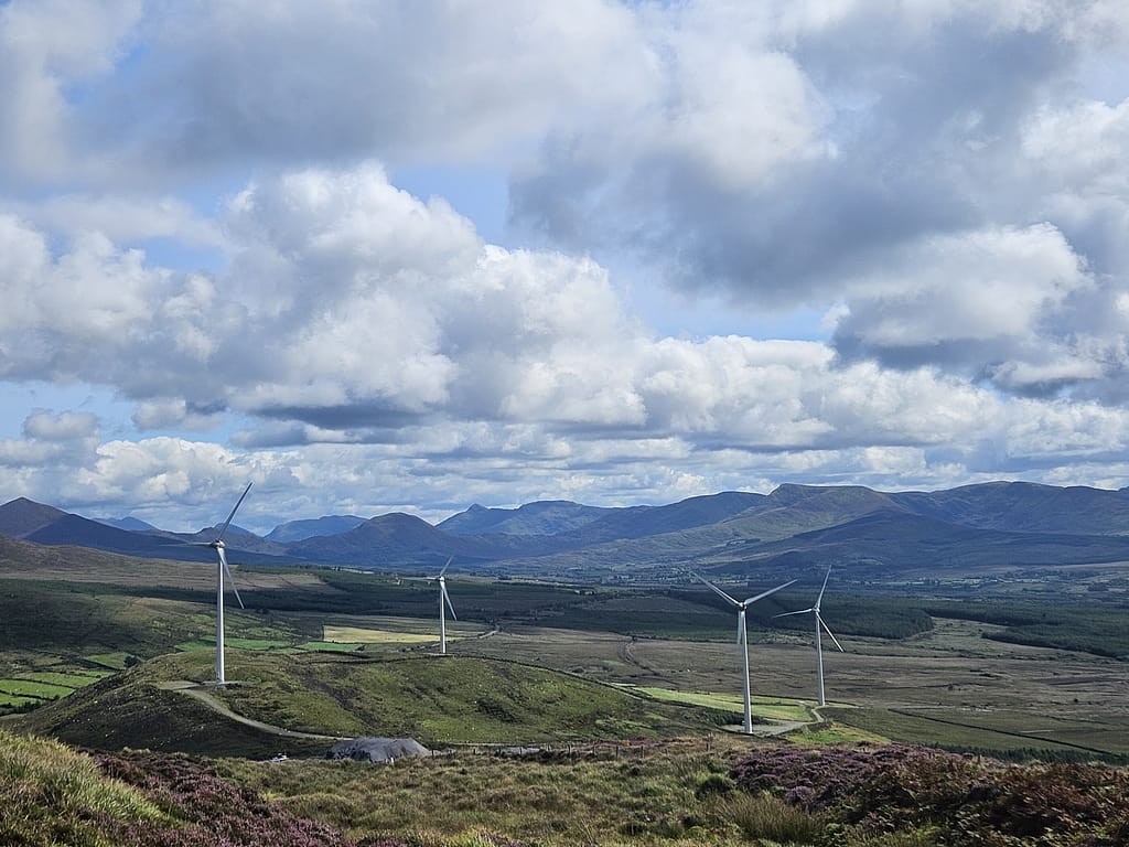 A vast mountain landscape, wind turbines in the foreground, green slopes and blue sky in the background.