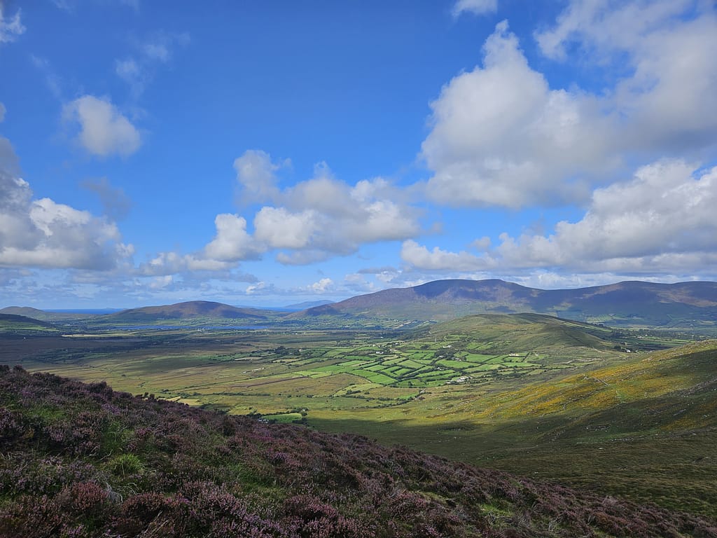 A view of a wide green valley, blue sky and white clouds with mountains in the background.