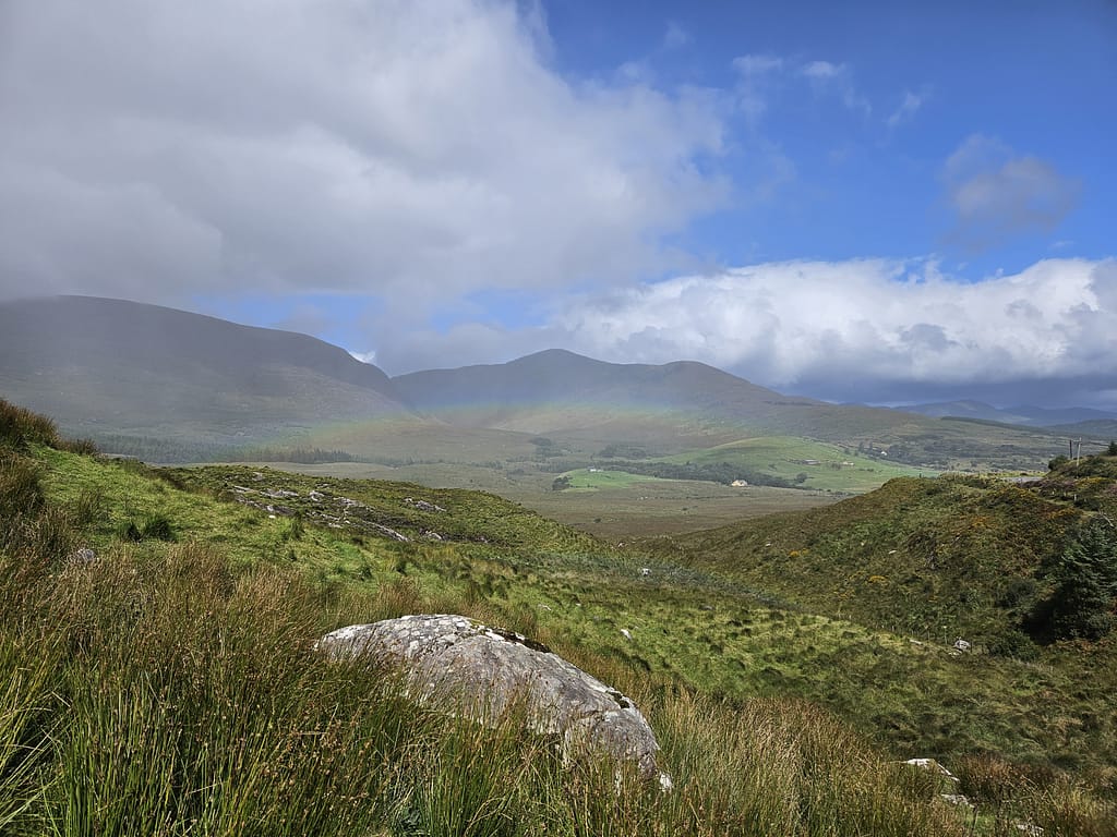 The landscape showcases the natural beauty of Ireland, with vast meadows, green mountains, and cloudy skies.