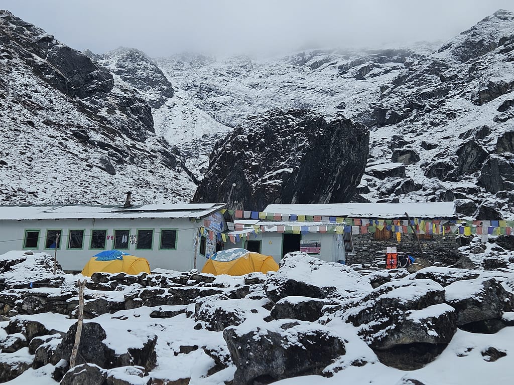 Karlı bir dağ manzarasıyla çevrili bir tea house, önünde yan yana dizilmiş sarı çadırlar ve dağ bayraklarıyla donatılmış bir alan.