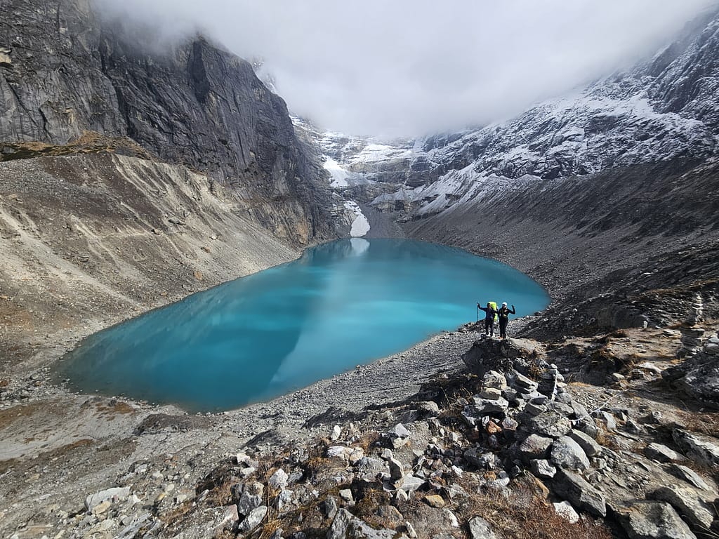 Himalayalar'da bir buzul gölü, çevresinde karla kaplı dağlar ve kayalarla birlikte yürüyüş yapan iki dağcı.