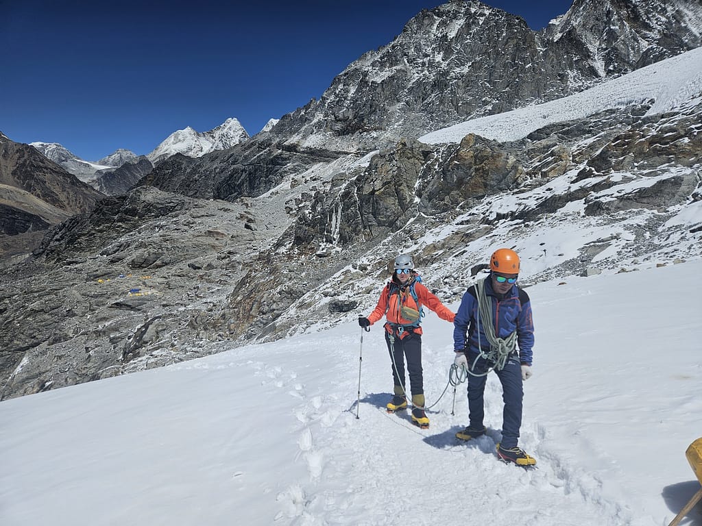İki dağcı, karlı bir dağ yörüngesinde ilerliyor. Arka planda yüksek dağlar ve mavi bir gökyüzü görülüyor. Dağcıların bir tanesi kırmızı bir ceket giymekte ve tahta baston kullanıyor, diğeri ise mavi bir giysi içinde tırmanmakta.