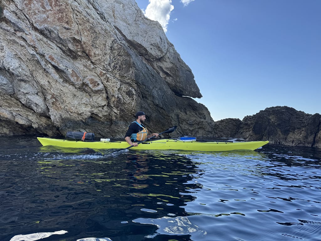 Bir adam, sarı bir deniz kayığında büyük kayalıklara yakın bir şekilde kürek çekiyor. Güneşli bir gün ve deniz durgun.