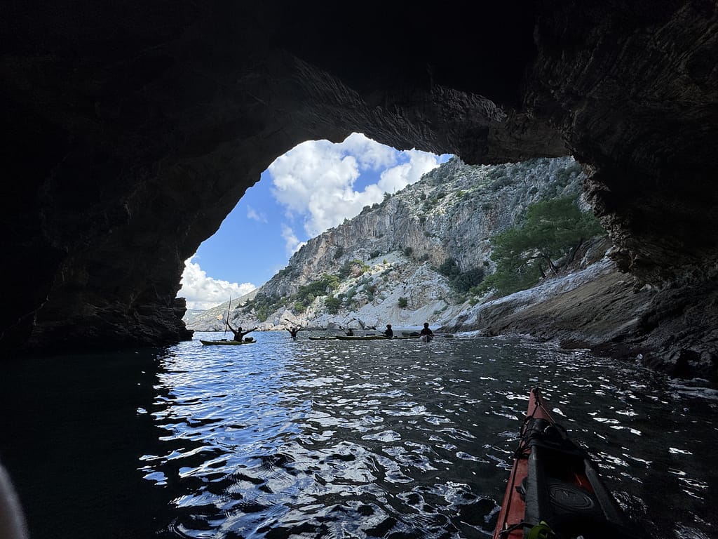 Bir kayıkçı grubu, büyük bir taş kemerin altından geçerken görünmektedir. Arkada deniz ve yeşil dağlık araziler yer almaktadır.