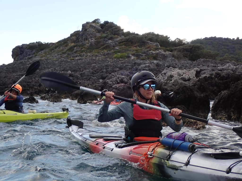 Deniz kayığı yapan katılımcılar, kayalık bir kıyı boyunca kürek çekiyor. Arkada kayalık detaylar ve doğal bir manzara görünmekte.