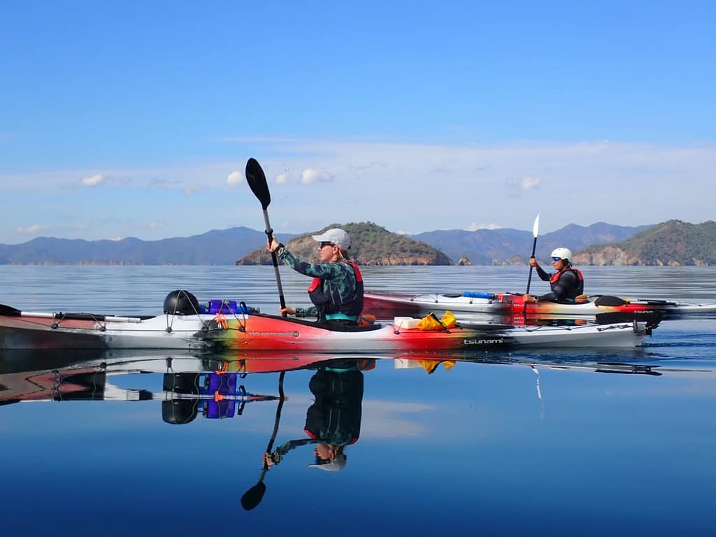 İki deniz kayağı kullanıcısı, sakin bir su yüzeyinde kürek çekerek ilerliyor.
