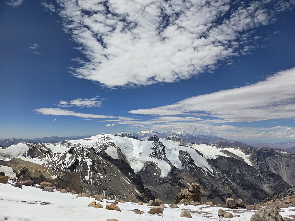 Beyaz karlarla kaplı dağ manzarası, mavi gökyüzünde bulutlarla birlikte görünüyor.