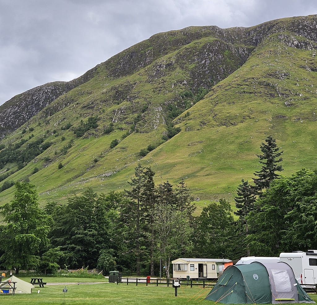 Il campeggio, adagiato su splendidi pendii verdeggianti, si trova ai piedi del Ben Nevis. Sullo sfondo si stagliano un cielo nuvoloso e le ripide pareti rocciose della montagna. Il campeggio offre tende da sole e roulotte.