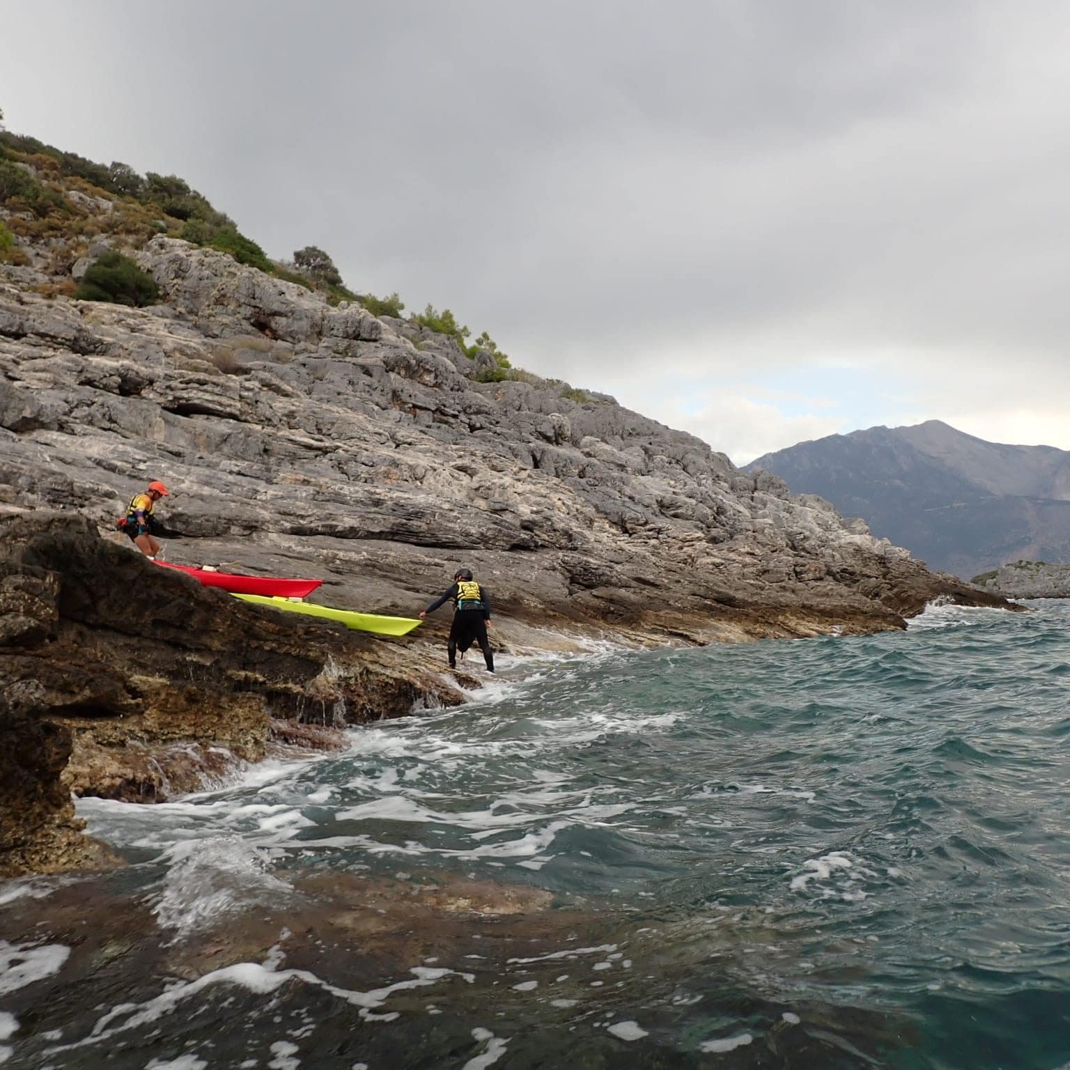 İki deniz kayağı, kayalık bir kıyıya yakın bir bölgede duruyor. Sağda bir kişi kayığı taşırken, solda başka bir kişinin kayığı suya indirirken görüldüğü bir sahne.