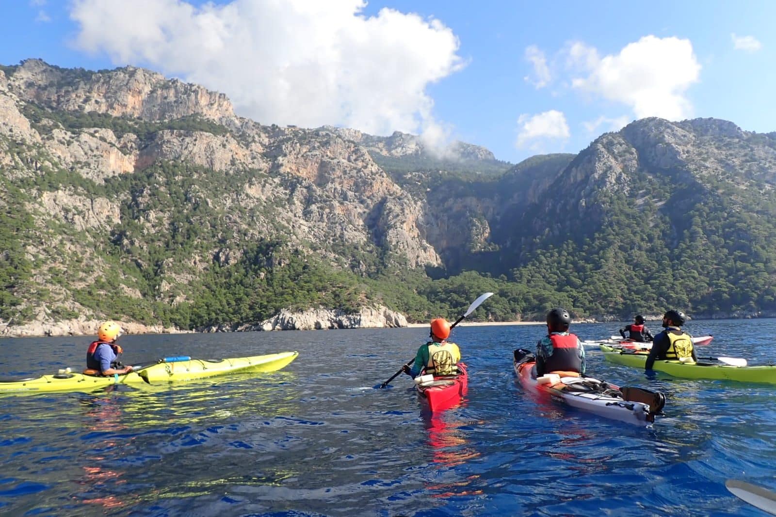 Deniz kayığı kullanan bir grup insan, dağlık ve ağaçlık bir kıyıya yakın sularda kaymakta.