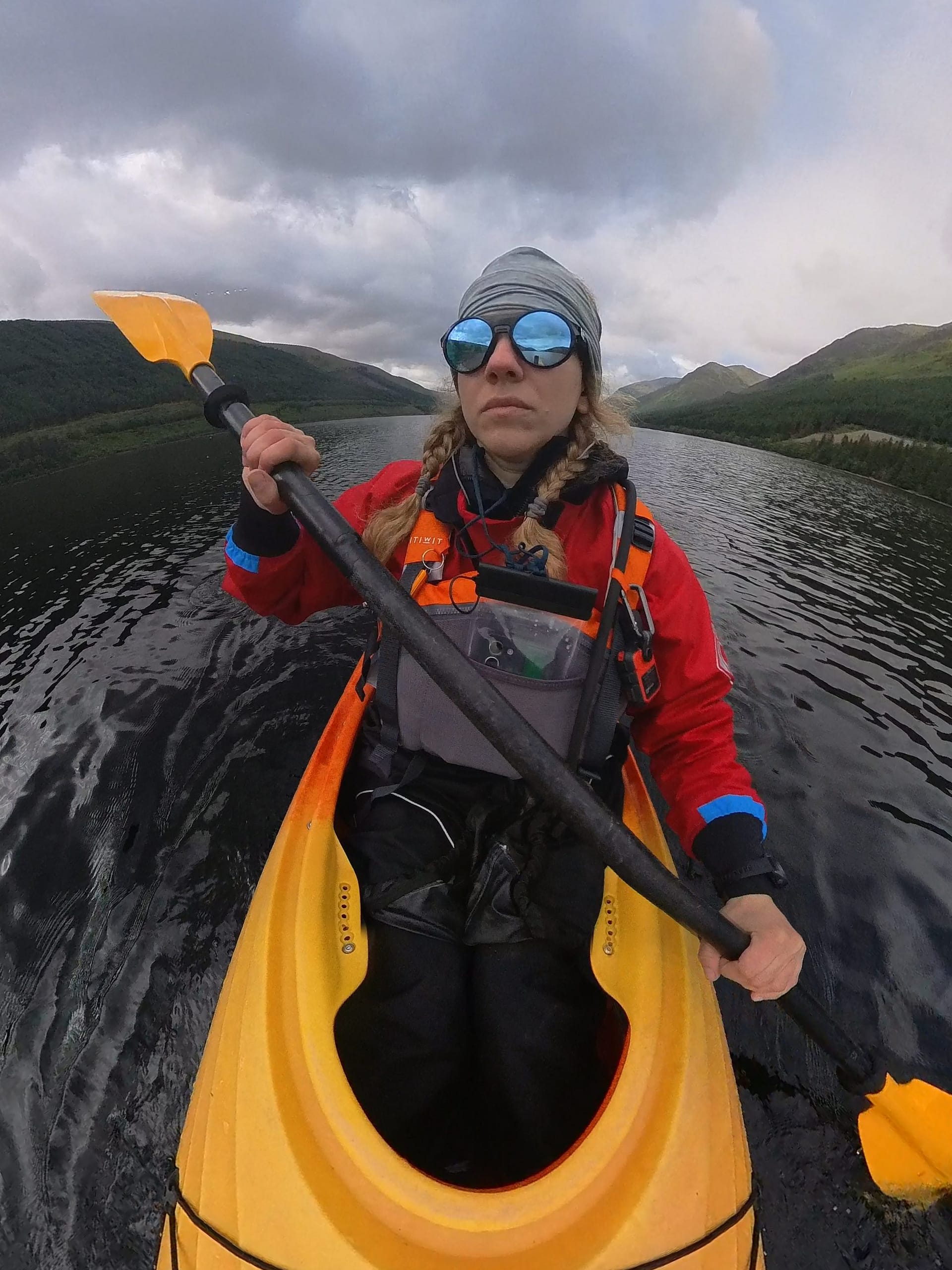 A person is seen kayaking on the water, wearing protective gear and sunglasses, with a green mountain landscape in the background.