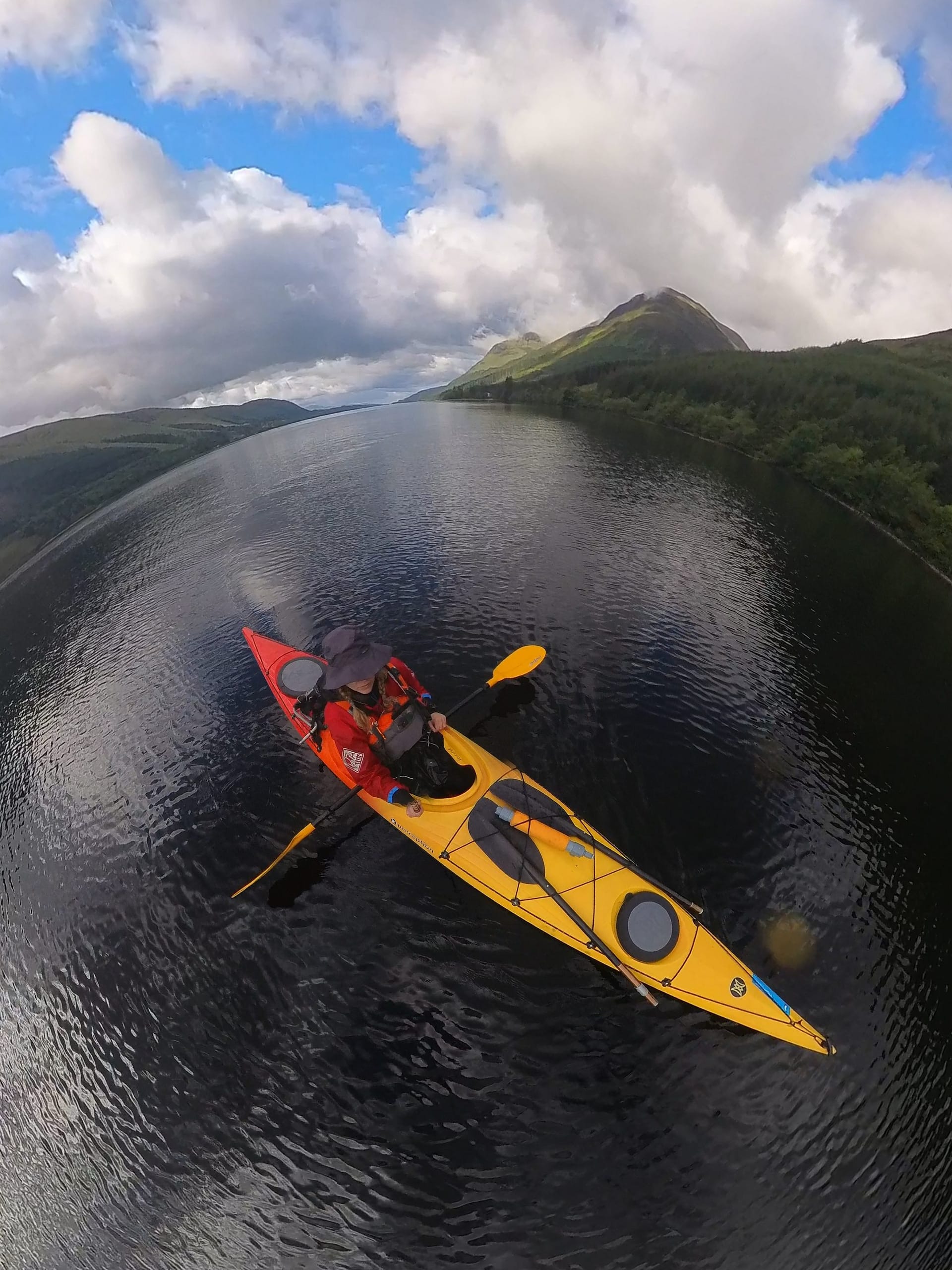 A person in a boat rows across a calm lake using a yellow kayak, with a backdrop of tall mountains and a cloudy sky.