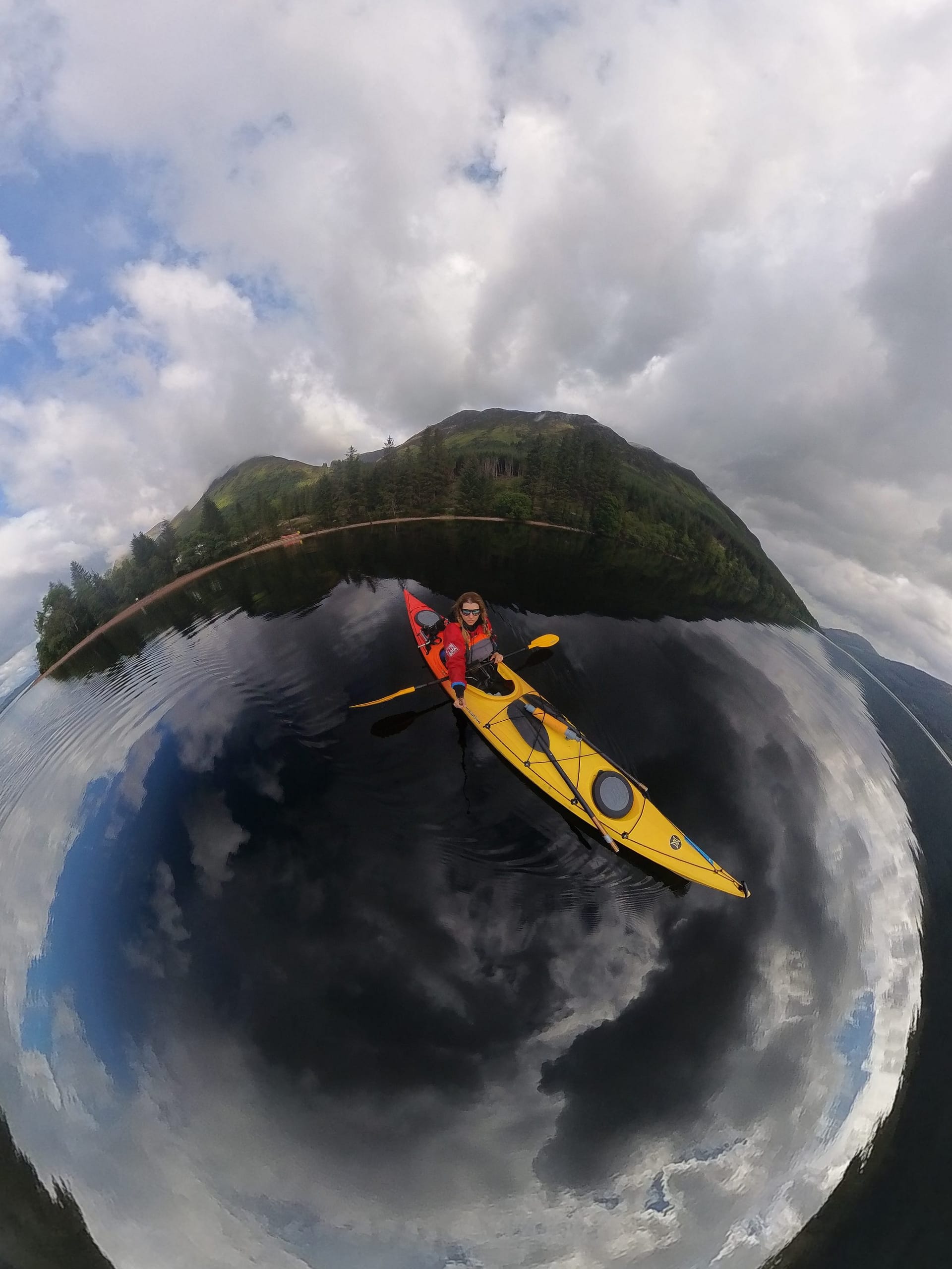 A person in a canoe boat is paddling on a loch, surrounded by lush green mountains and clouds in the sky.