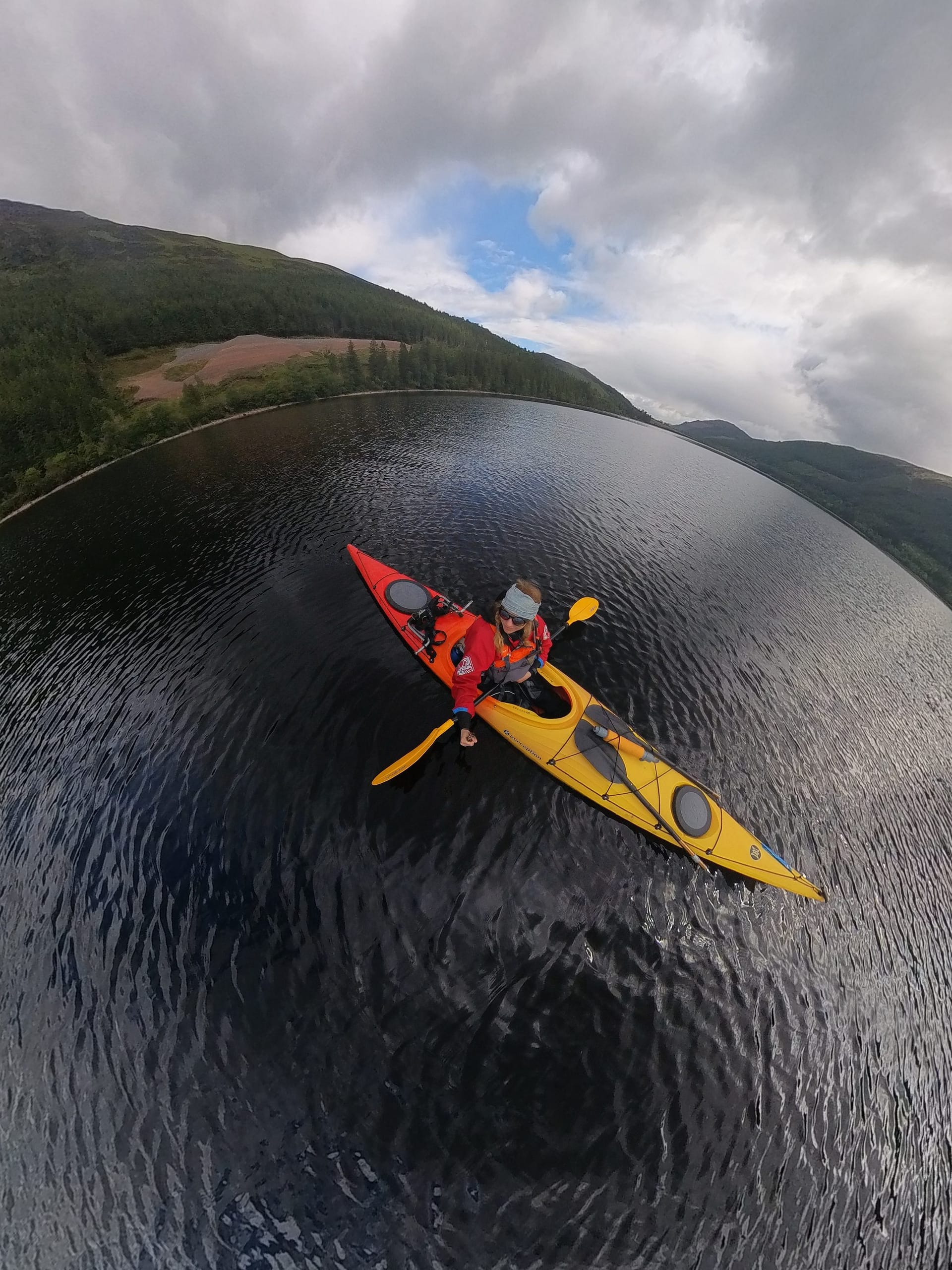 A person is rowing a yellow and red kayak across the water, surrounded by green mountains and clouds in the sky.