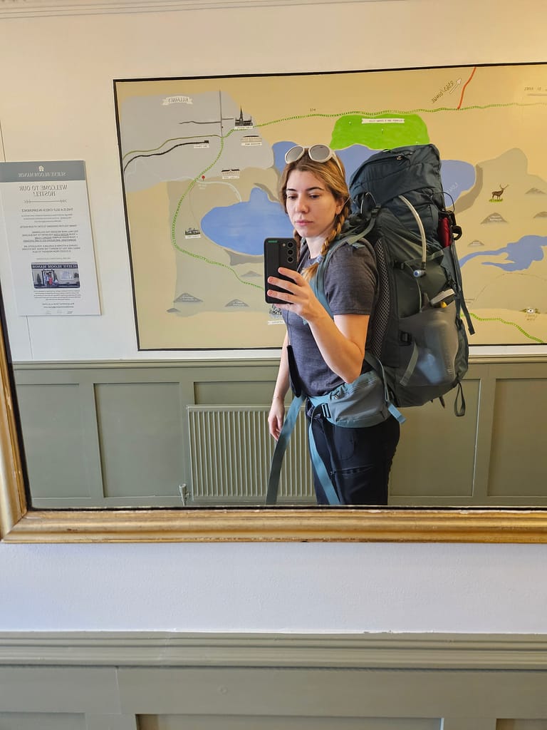 A woman takes a selfie in the mirror, wearing a large backpack. A nature map is visible in the background.