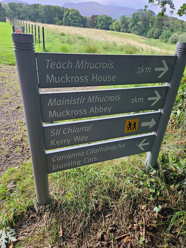 Road sign pointing towards Muckross House and Kerry Way, surrounded by green nature scenery.