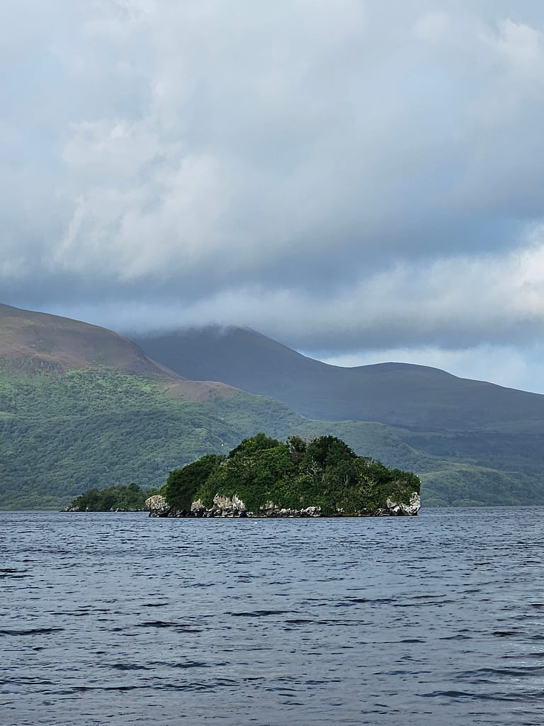 The image of a small island and its surrounding waters merges with the green slopes of distant mountains and cloudy skies.