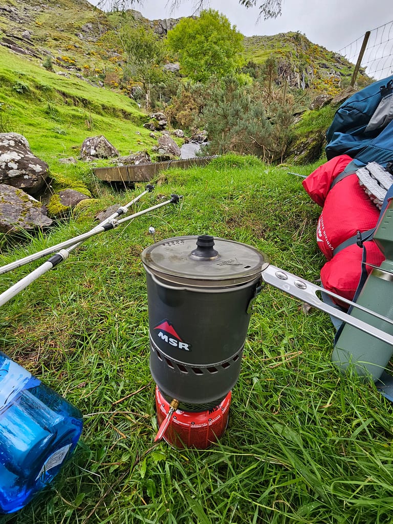 Water boils on a stove sitting in a grassy area, with green vegetation and bags surrounding it.
