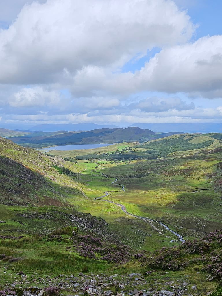 A sweeping valley, lush green pastures, and a gently rippling lake. In the distance, silhouetted mountains and a cloud-covered sky.