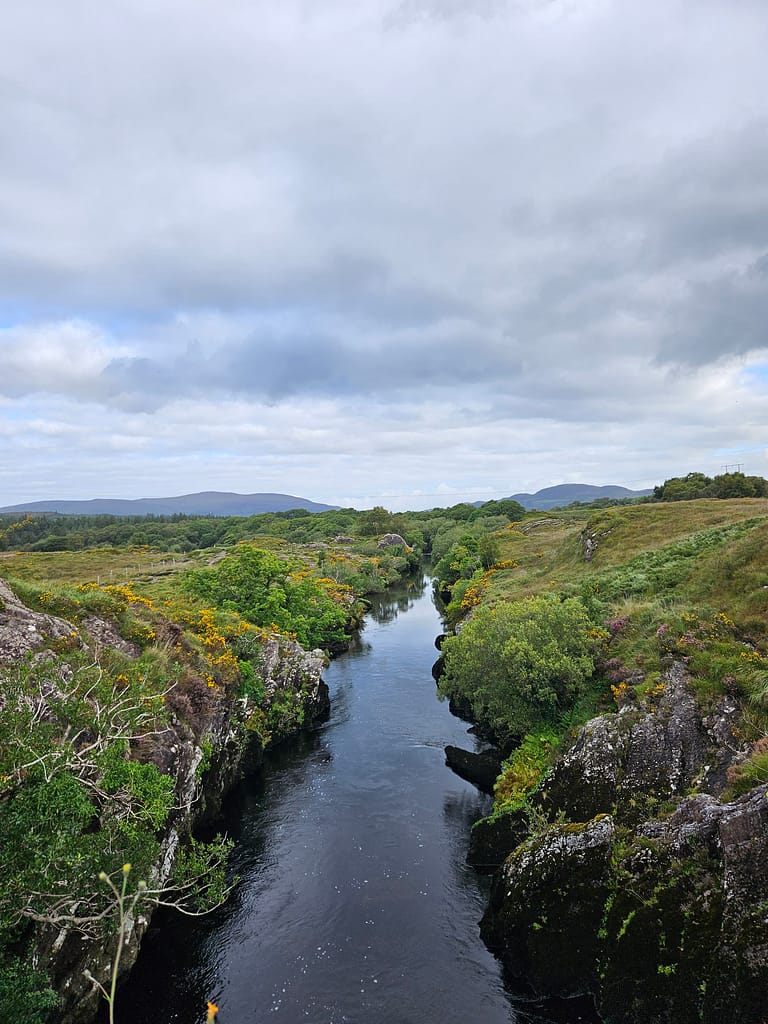 A river flowing through a natural valley landscape is seen with surrounding green vegetation and a cloudy sky.