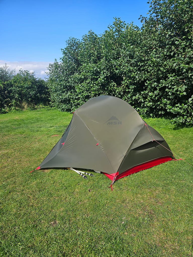 The MSR branded tent, set up in a camping area, is positioned above the ground on a green grass area.