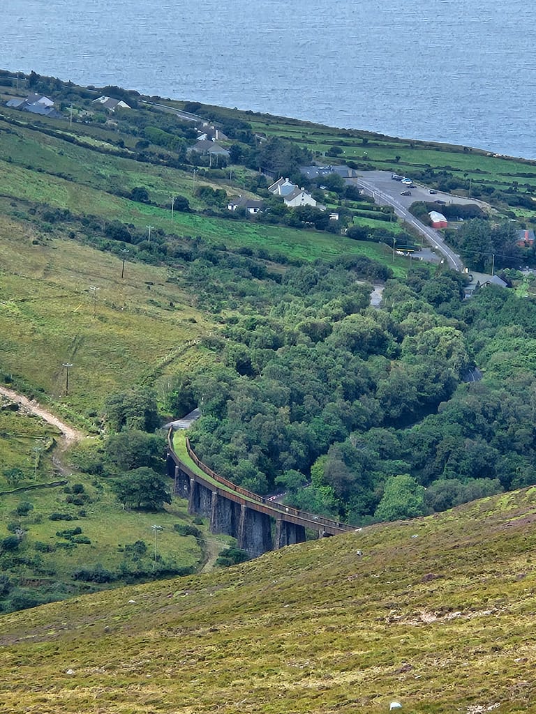 The image shows a landscape of lush green fields and the sea. In the distance, a road and a few houses surrounded by trees are visible. A viaduct sits on a green hillside in the foreground.