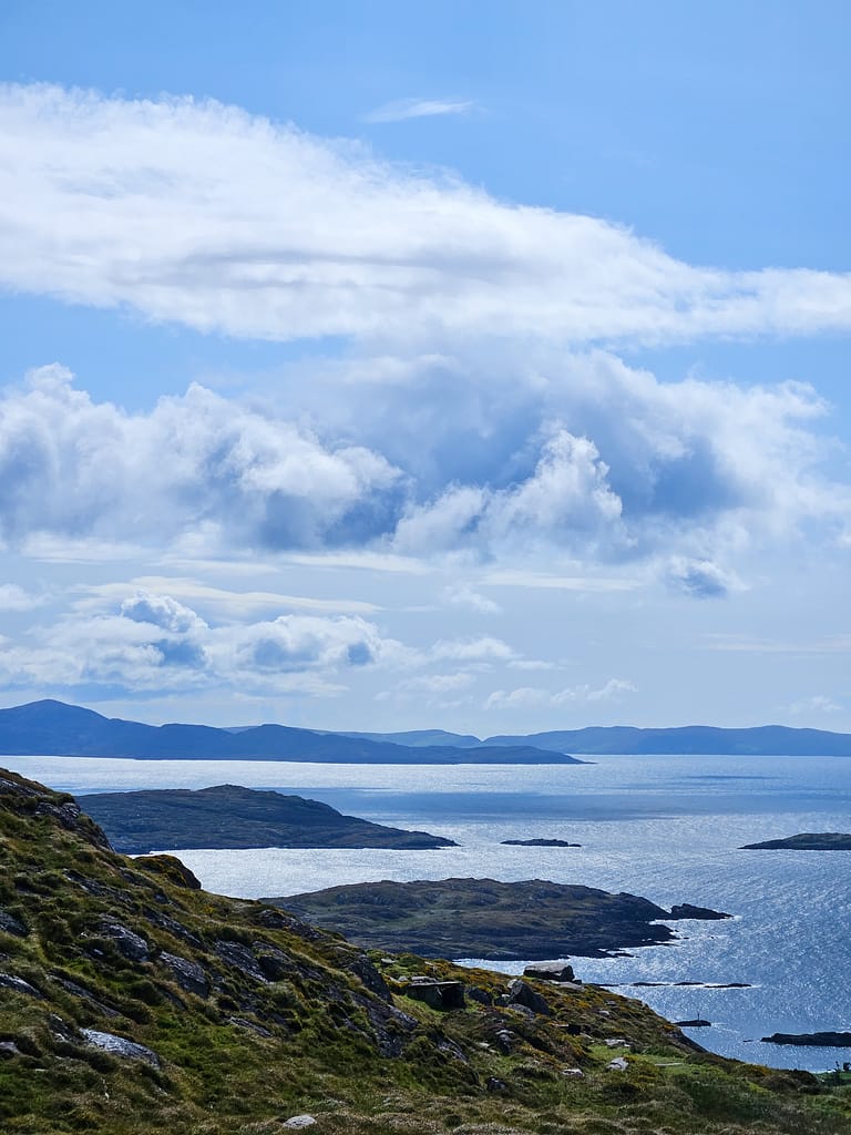 The clear blue sky and cloud-covered landscape merge with the sea and islands – an Atlantic landscape where green hills meet the sea, reflecting the natural beauty of Ireland.
