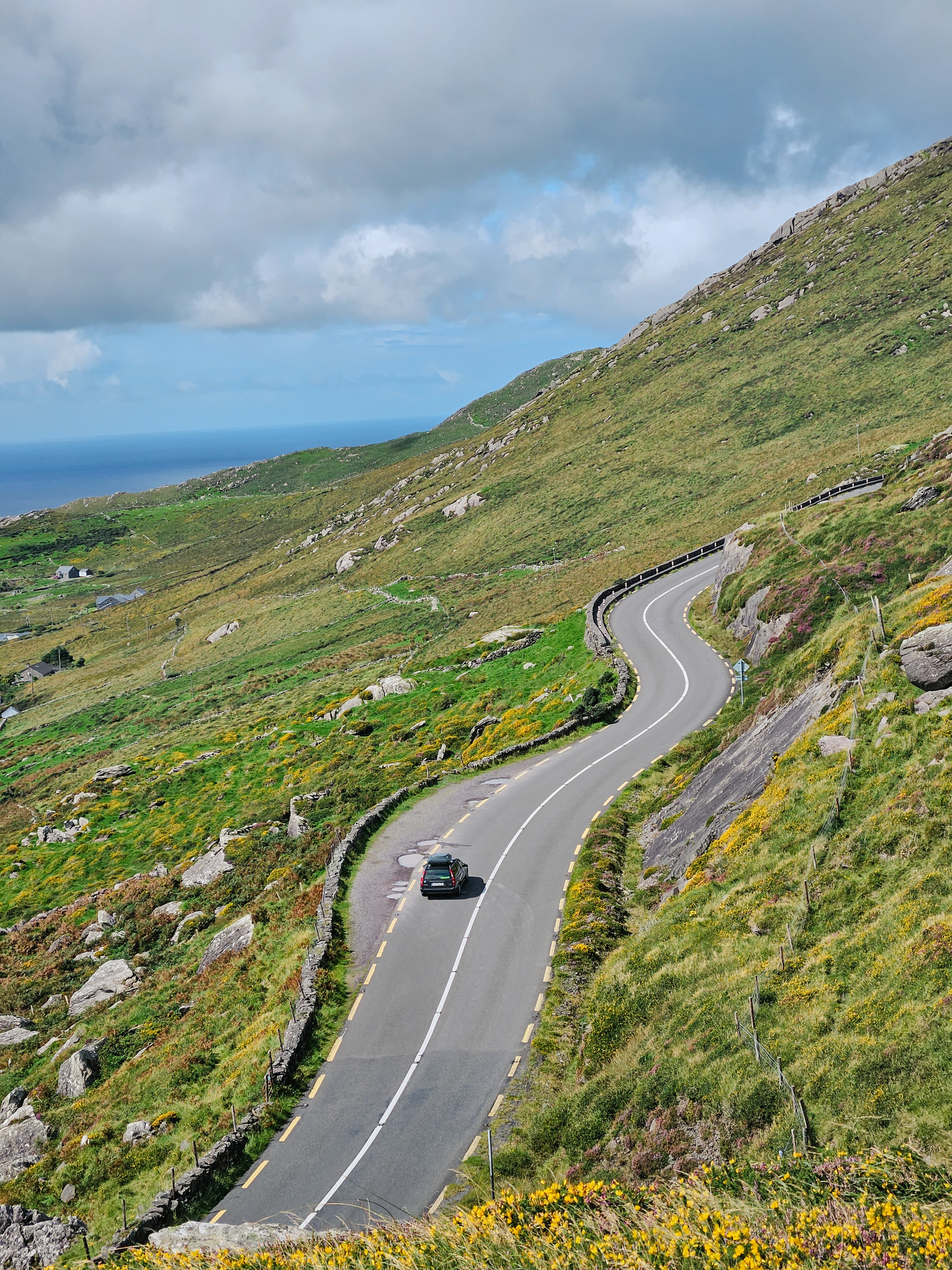 A winding road winds through green hills and, in the distance, sea views with cloudy skies, reflecting the natural beauty of Ireland.