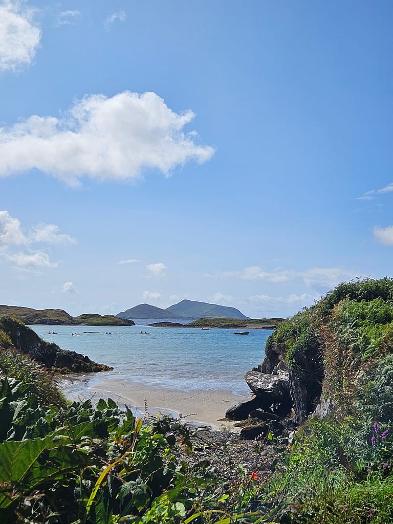 A beach scene with turquoise water and boats floating on it. In the background are green hills and a clear blue sky.