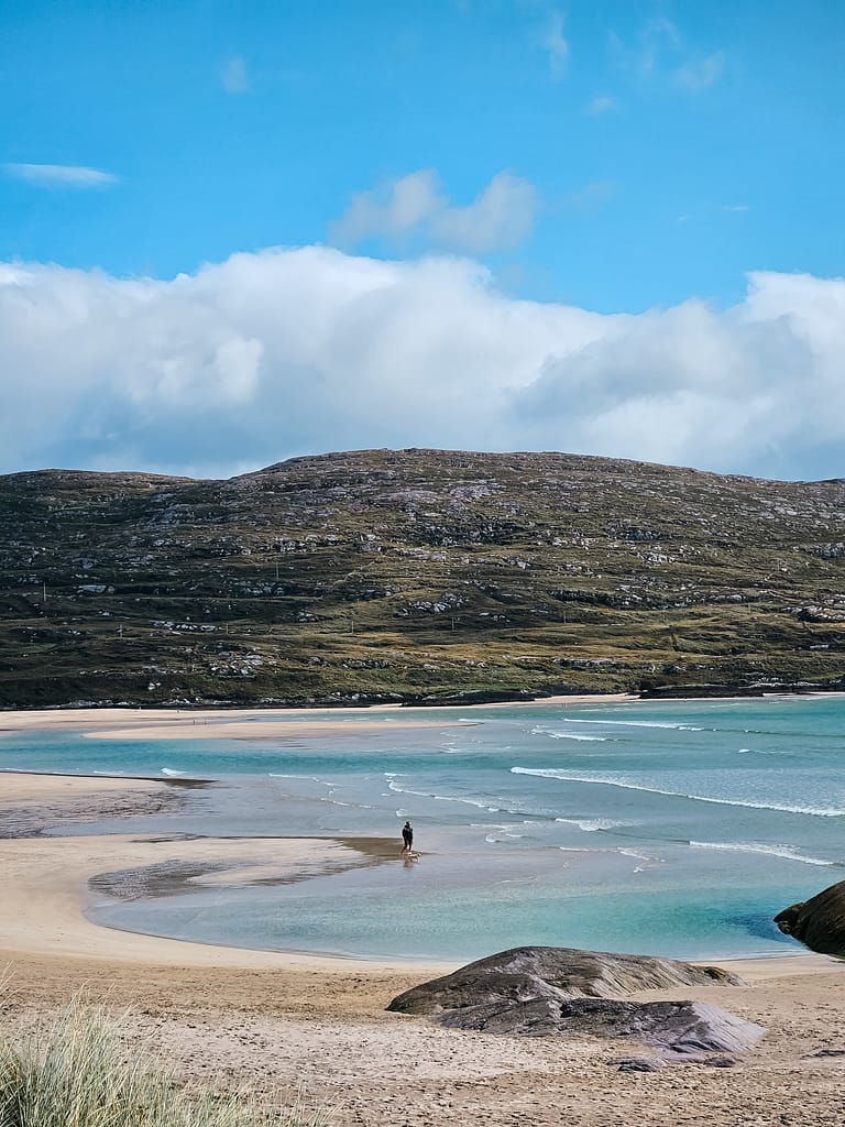 Coastal landscape with green hills and clear blue water and sandy beach with a man walking along the shore.