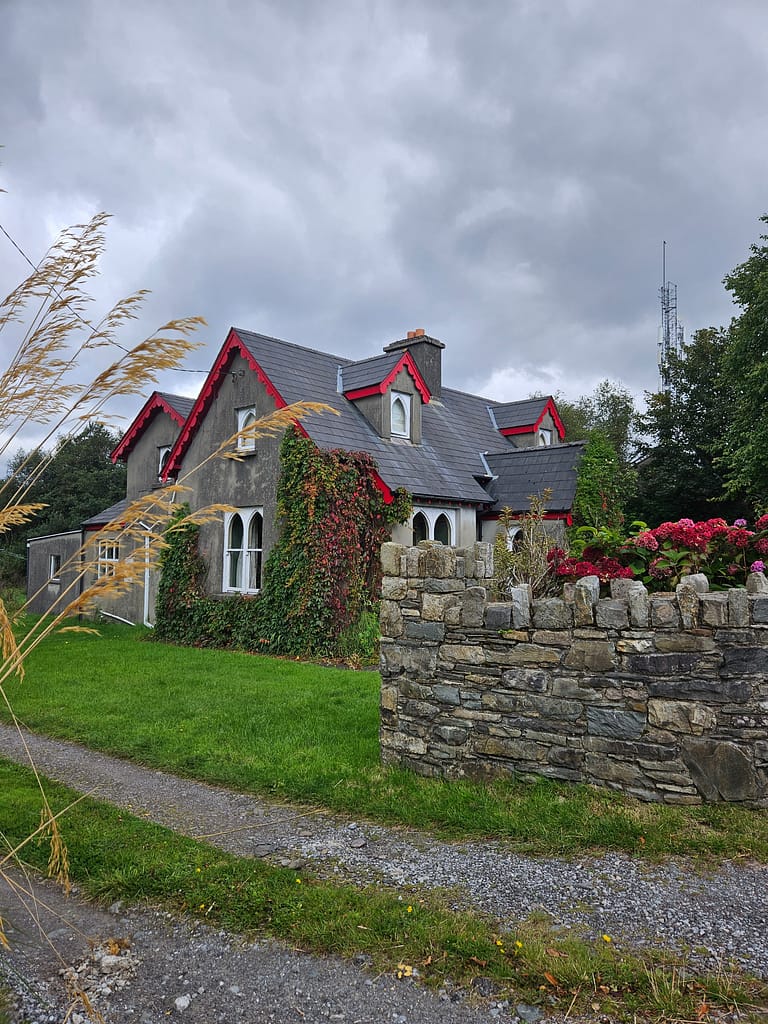 The front of a house is surrounded by a red roof and windows, surrounded by greenery and a stone wall.