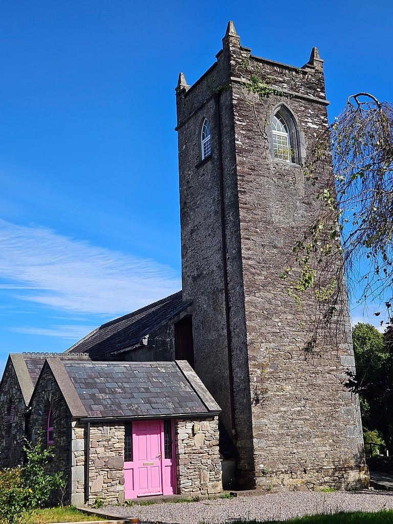 A stone church with a tall bell tower and a pink door. A smaller building is visible next to it, surrounded by a cloudy blue sky.
