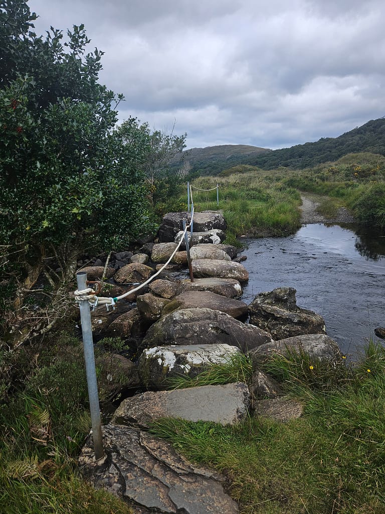 A stone bridge in a natural setting, topped by an iron bolt, with a green hillscape in the background. The weather is overcast and cloudy.