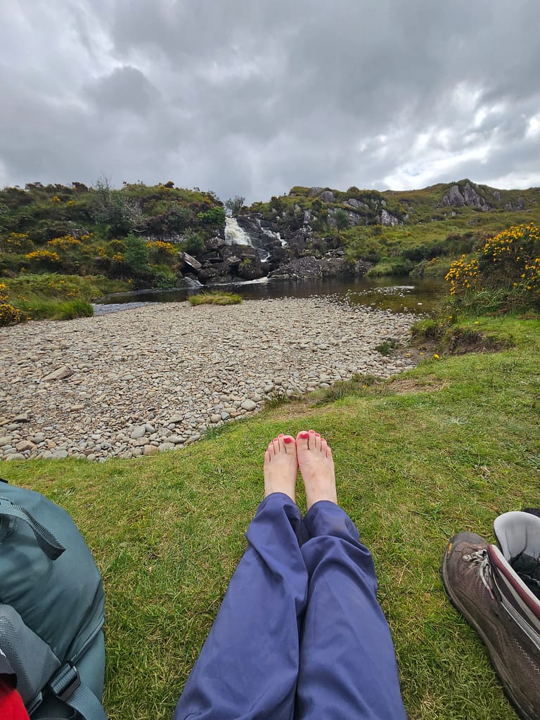 The feet of a person sitting in a natural landscape are visible; in the background there is a small waterfall and rocks among green plants.
