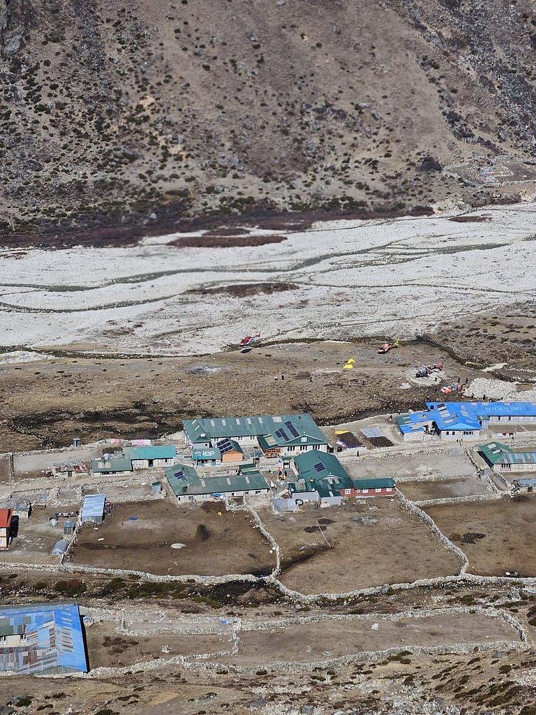 Himalaya bölgesinde bulunan bir yerleşim alanının uzaktan görünümü. Yeşil çatıları olan binalar ve çevresinde tarımsal veya doğal alanlar görünmektedir.