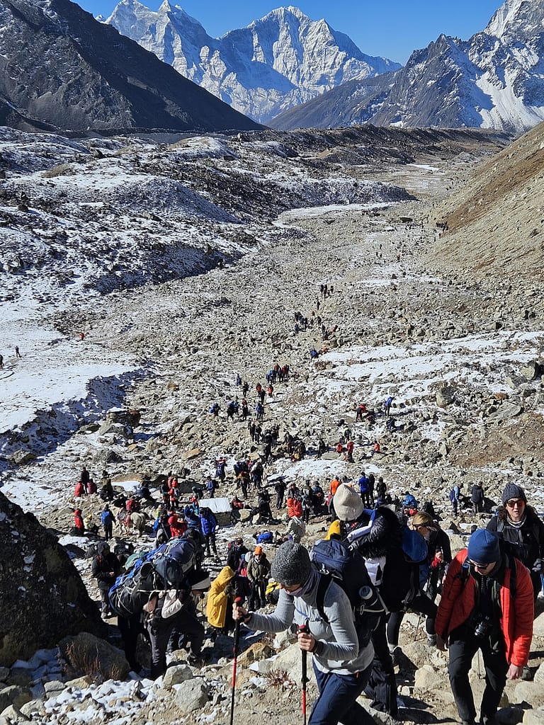 Bir grup dağcı, karla kaplı dağlık arazide Everest Base Camp yürüyüşü için ilerliyor. Arkada yüksek dağlar ve mavi gökyüzü görünmekte.