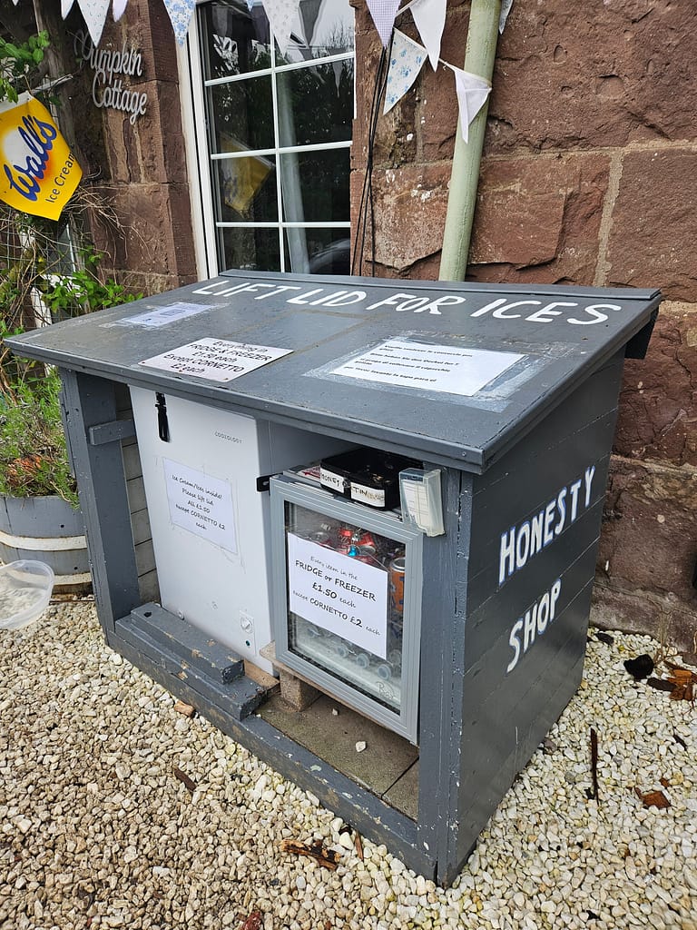 Bir honesty shop (güven dükkânı) görünümü, açık mavi bir tezgah üzerindeki dondurma ve içecek satışıyla birlikte dükkanın yanında.