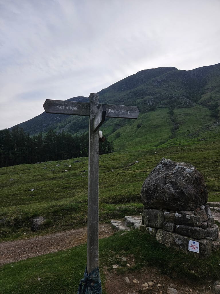 Un cartello indica la direzione di Ben Nevis e Achintree, con un paesaggio montuoso verde sullo sfondo.