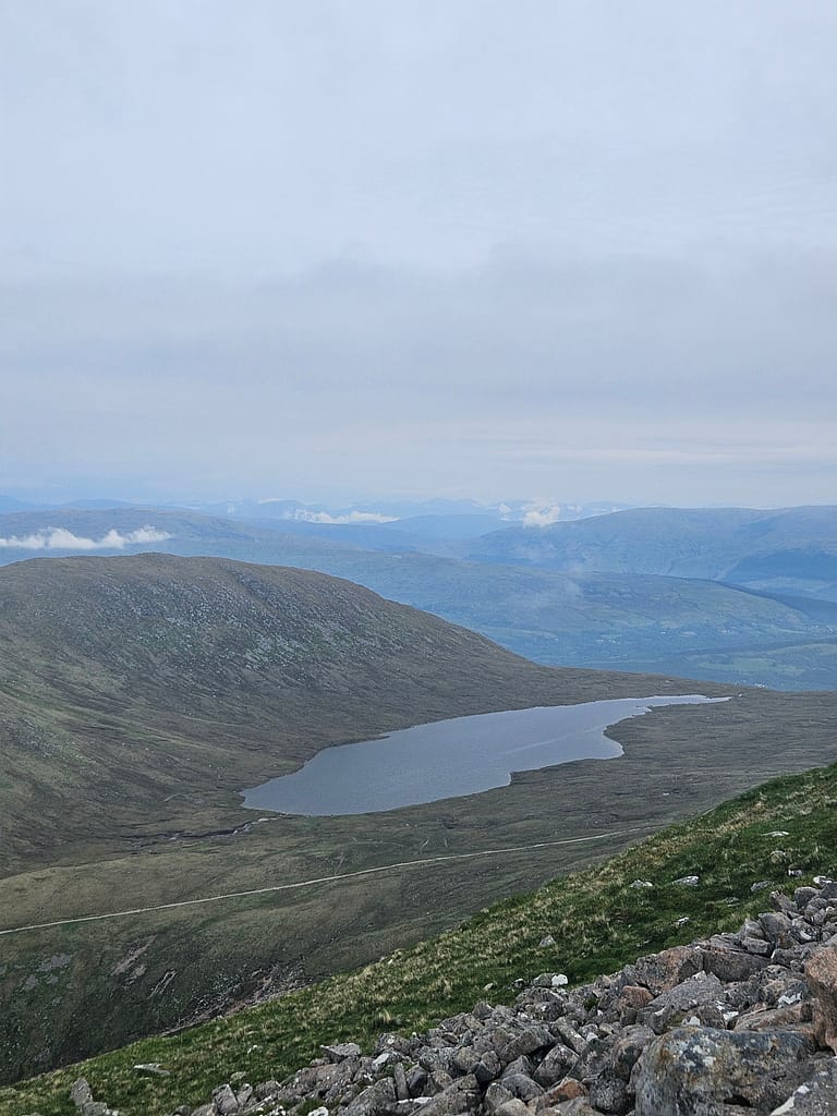 Una vista grandangolare del paesaggio montano dalla cima del Ben Nevis, con un lago in primo piano e catene montuose ondulate sullo sfondo.