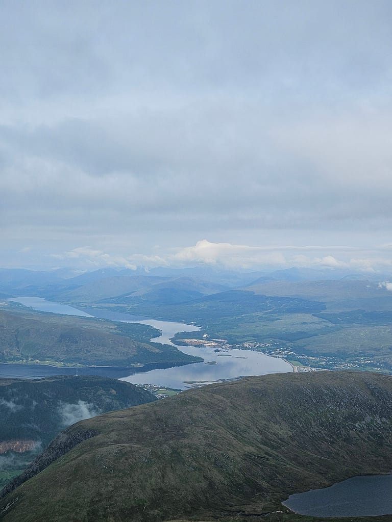 La vista dalla cima del Ben Nevis offre una vista spettacolare sul lago e sulle montagne. Il cielo è nuvoloso, con verdi colline e montagne in lontananza visibili.
